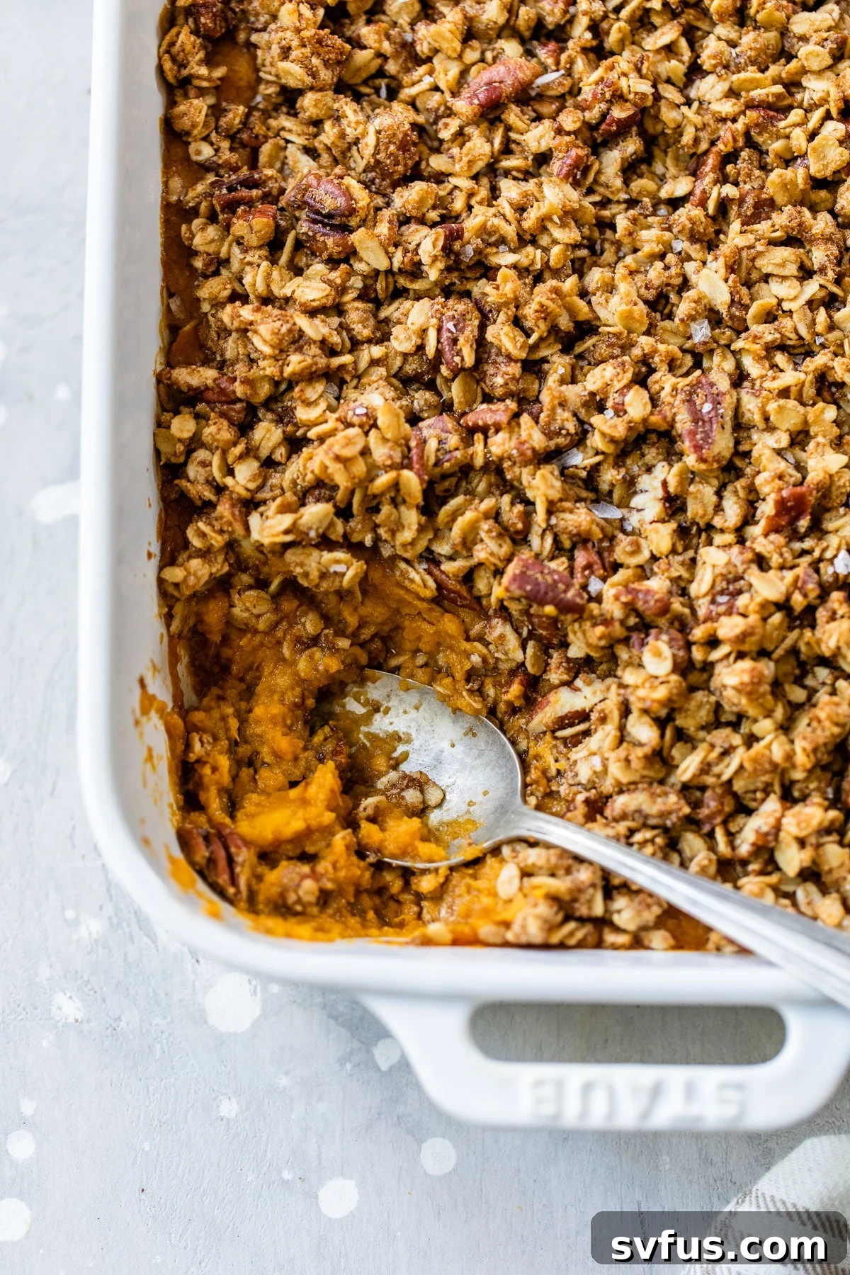A close-up shot of a golden-brown sweet potato casserole with a serving spoon, showing the crunchy pecan and oat topping and creamy interior.