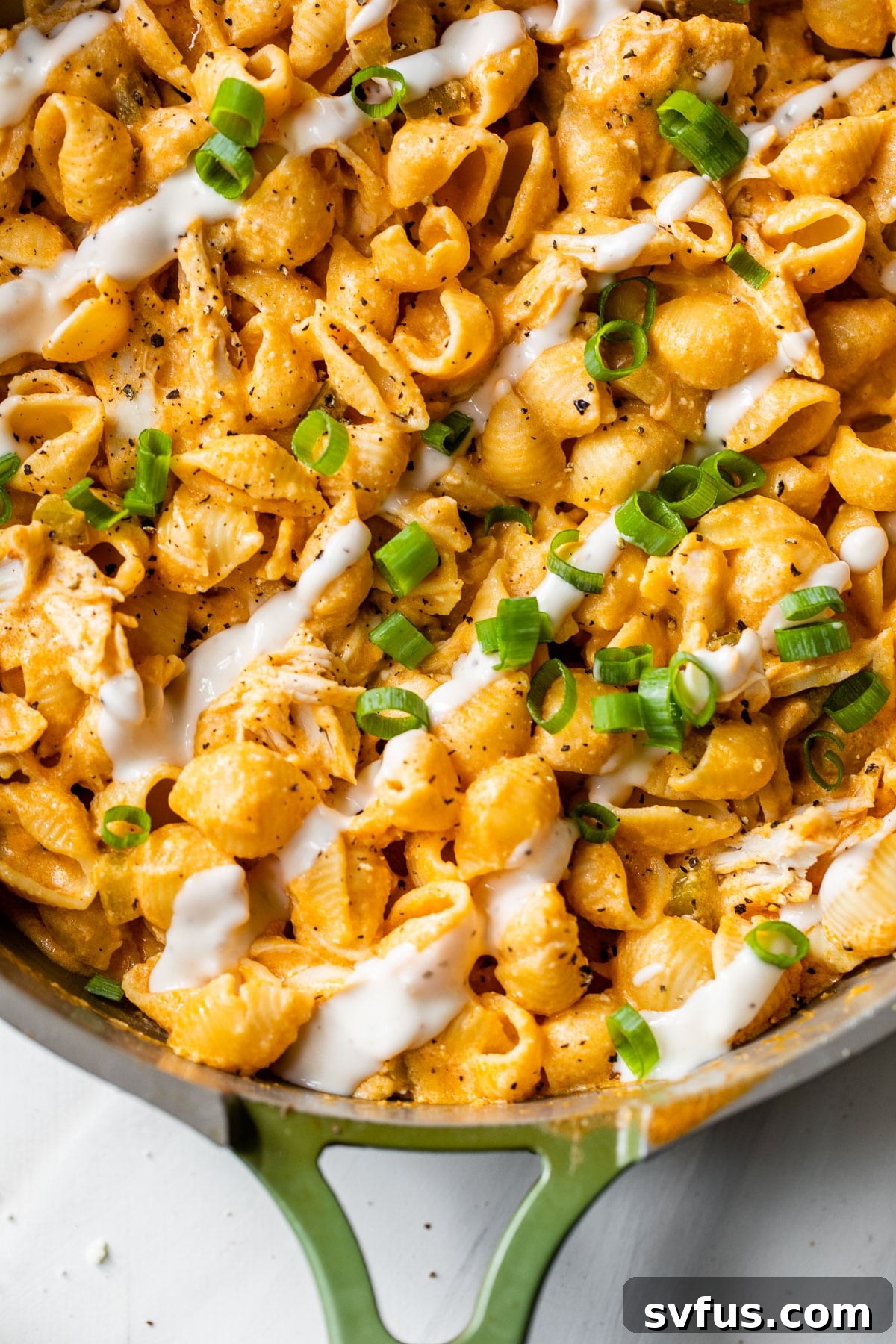 Close-up of buffalo chicken pasta in a skillet, topped with a drizzle of ranch dressing and fresh green onions.