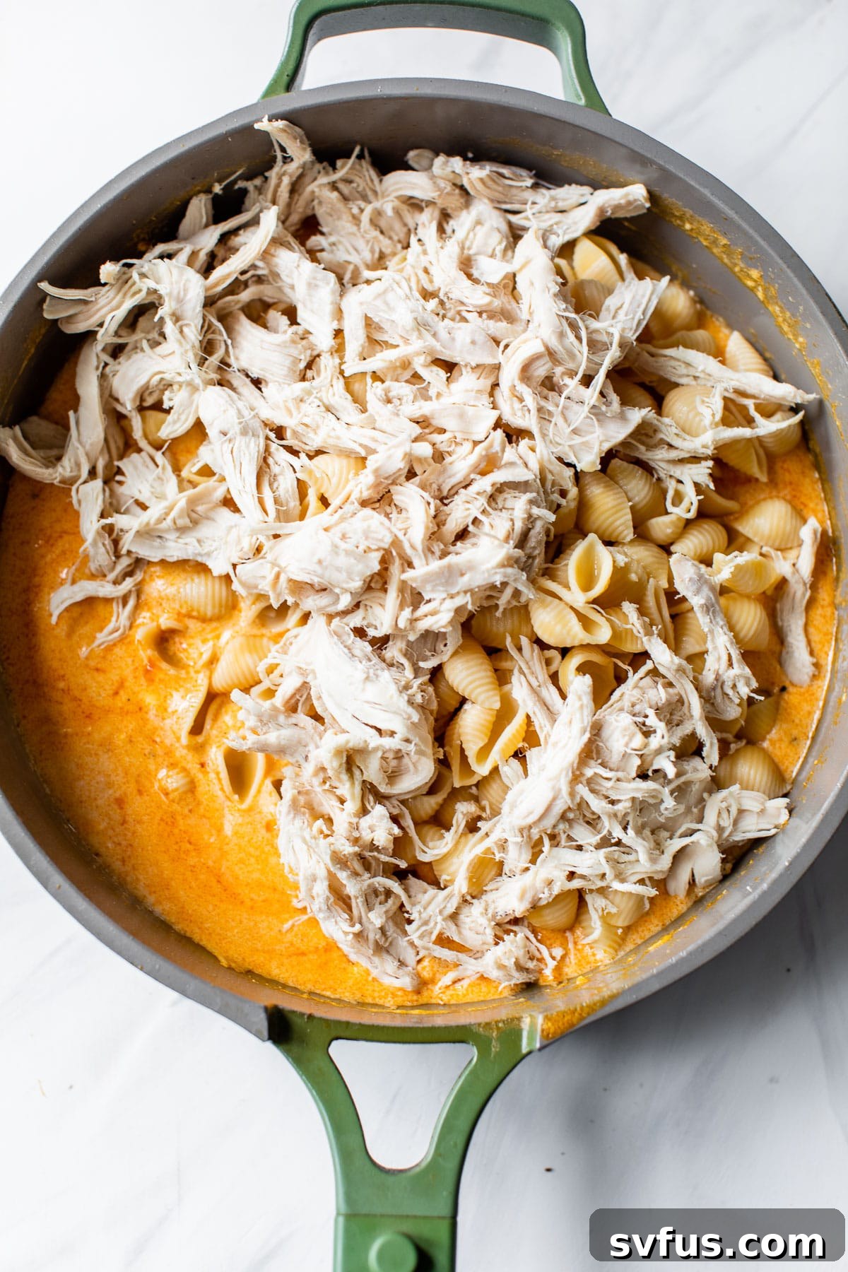 Cooked pasta and shredded chicken being tossed in the creamy buffalo sauce within a large skillet, showing the full mixture.