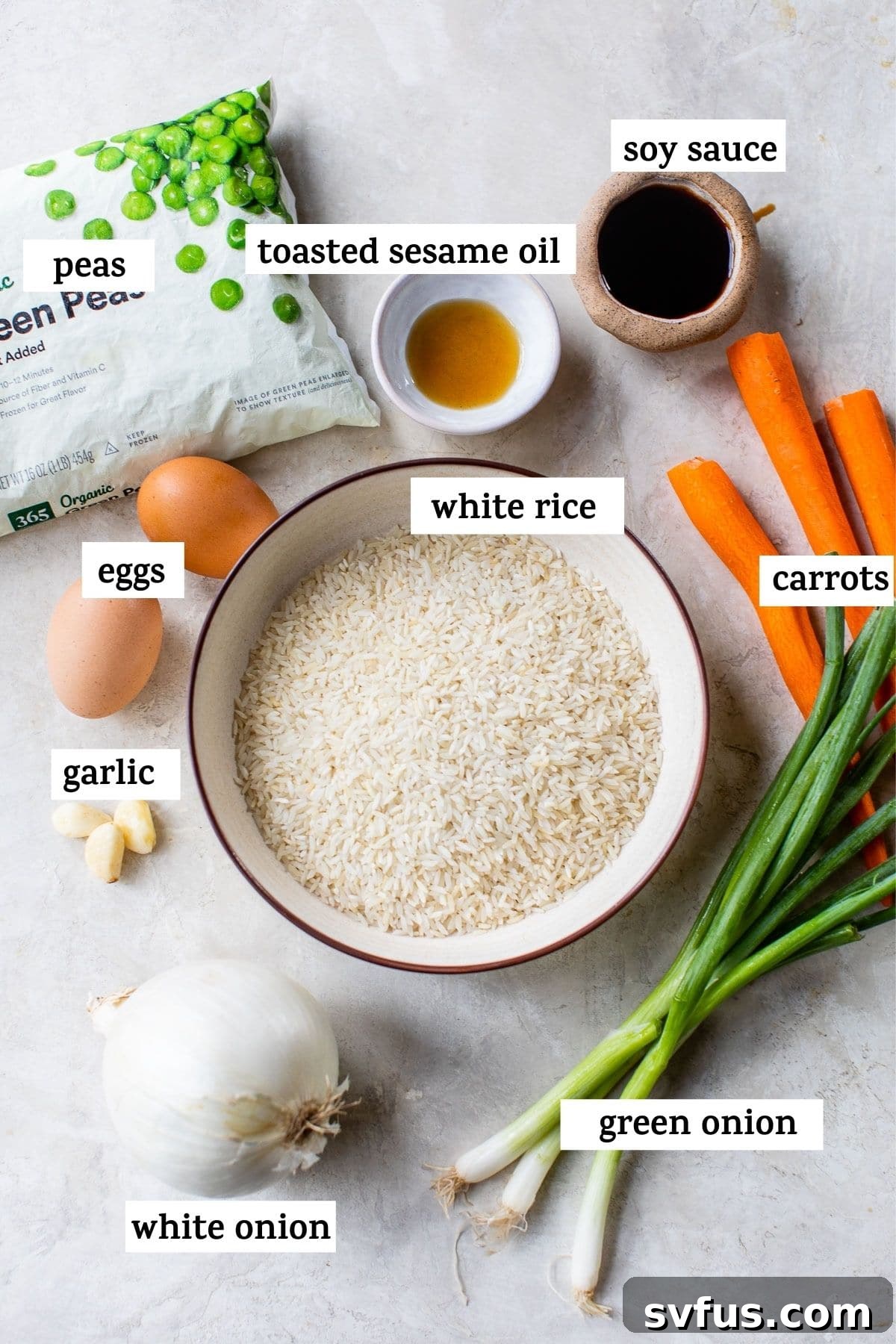 A flat lay of fresh ingredients arranged to make fried rice, including rice, carrots, peas, onion, garlic, green onions, eggs, butter, sesame oil, and soy sauce.