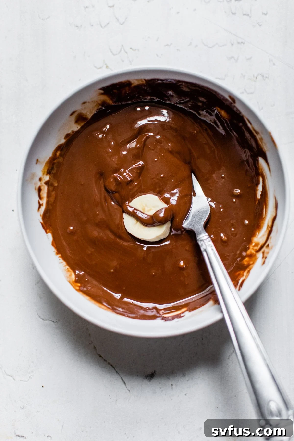 A banana slice being gently dipped into a bowl of rich, melted chocolate and peanut butter mixture, showcasing the smooth coating process.
