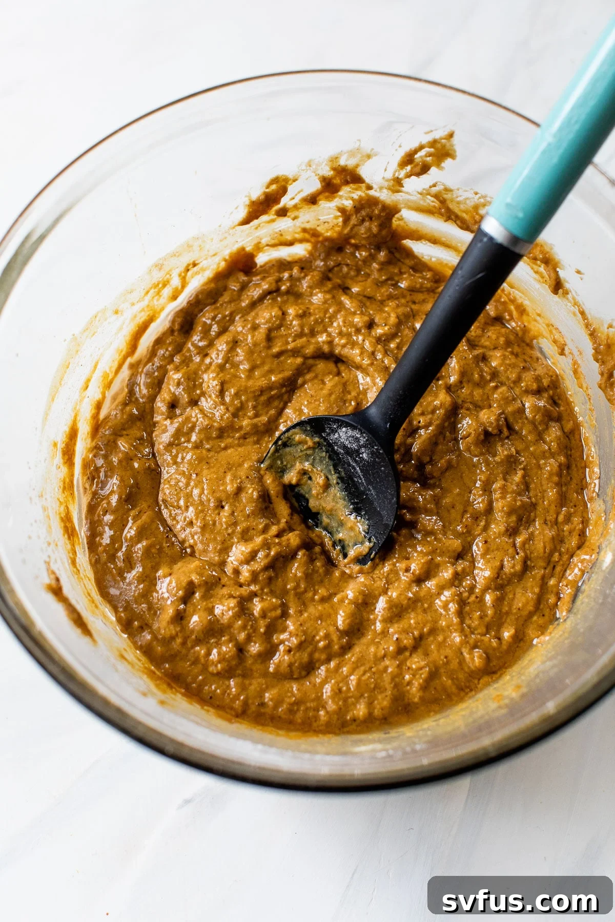 Muffin batter, still slightly lumpy, being gently mixed in a glass bowl with a rubber spatula, indicating it's ready for baking.