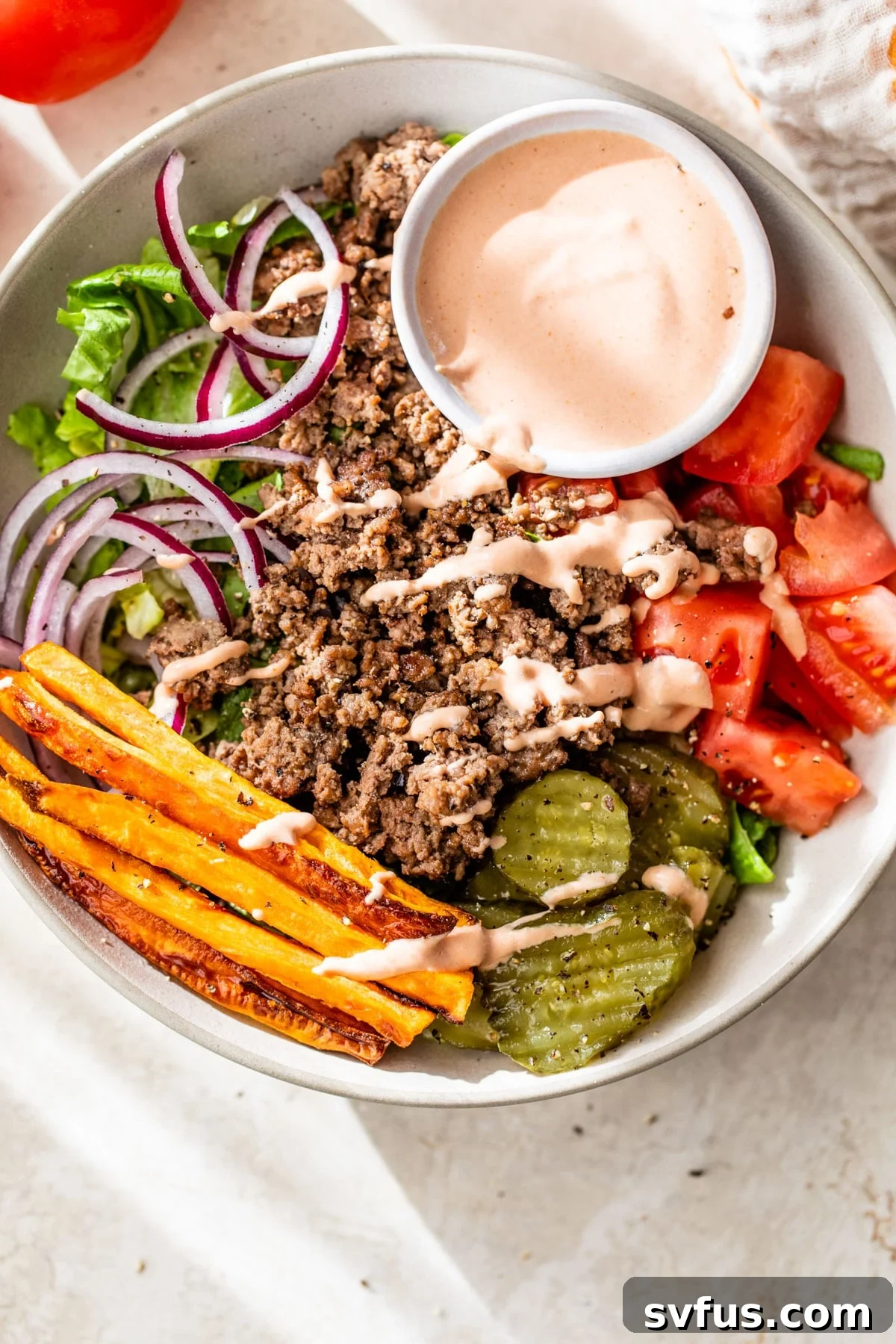 A vibrant burger bowl featuring crisp romaine lettuce, seasoned ground beef, roasted sweet potatoes, fresh tomatoes, and tangy pickles, all drizzled with a delicious sauce.