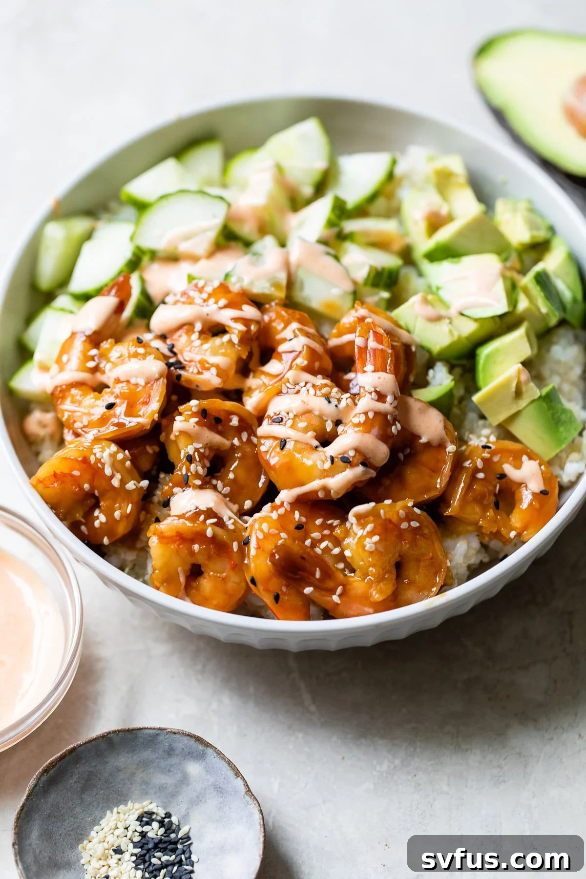 A vibrant sushi bowl featuring seasoned rice, tender cooked shrimp, fresh cucumber, and creamy avocado, drizzled with a spicy mayo.