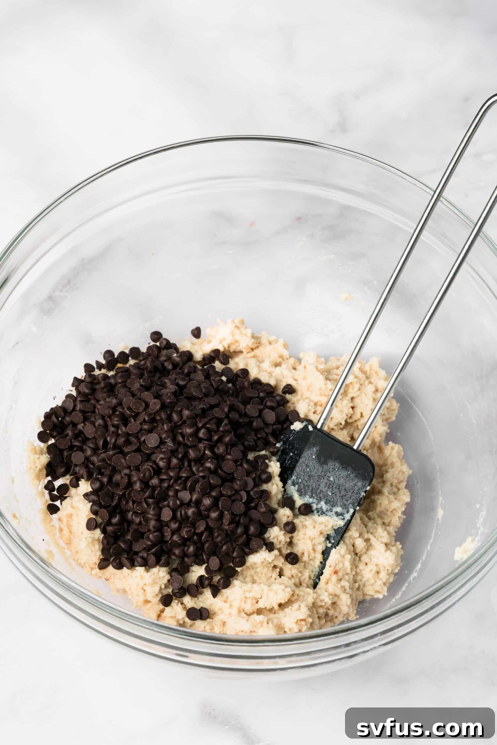 Side view of a black spatula mixing chocolate chips into cottage cheese cookie dough in a glass bowl.