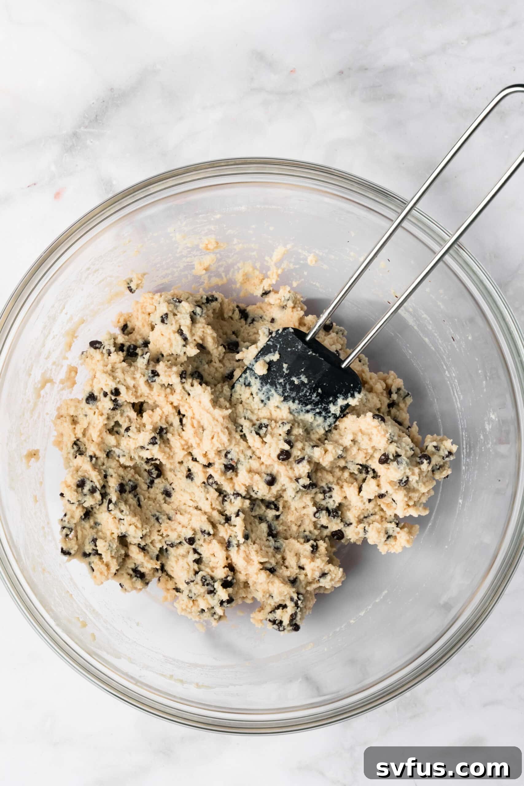 Overhead view of a black spatula mixing chocolate chip cottage cheese cookie dough in a glass bowl.