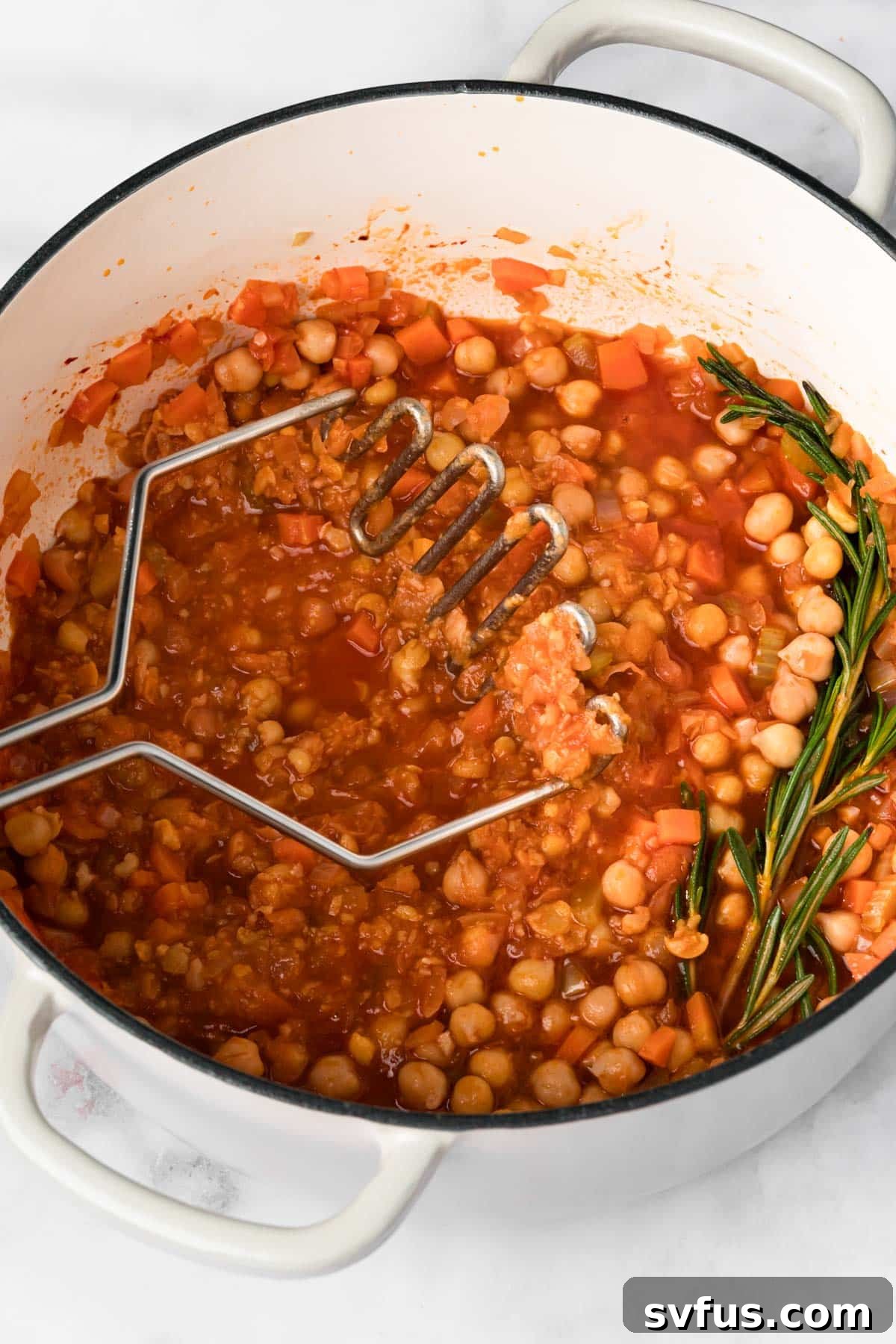 Overhead view of a potato masher pressing down on chickpeas in a pot of simmering Pasta e Ceci, demonstrating the mashing technique.