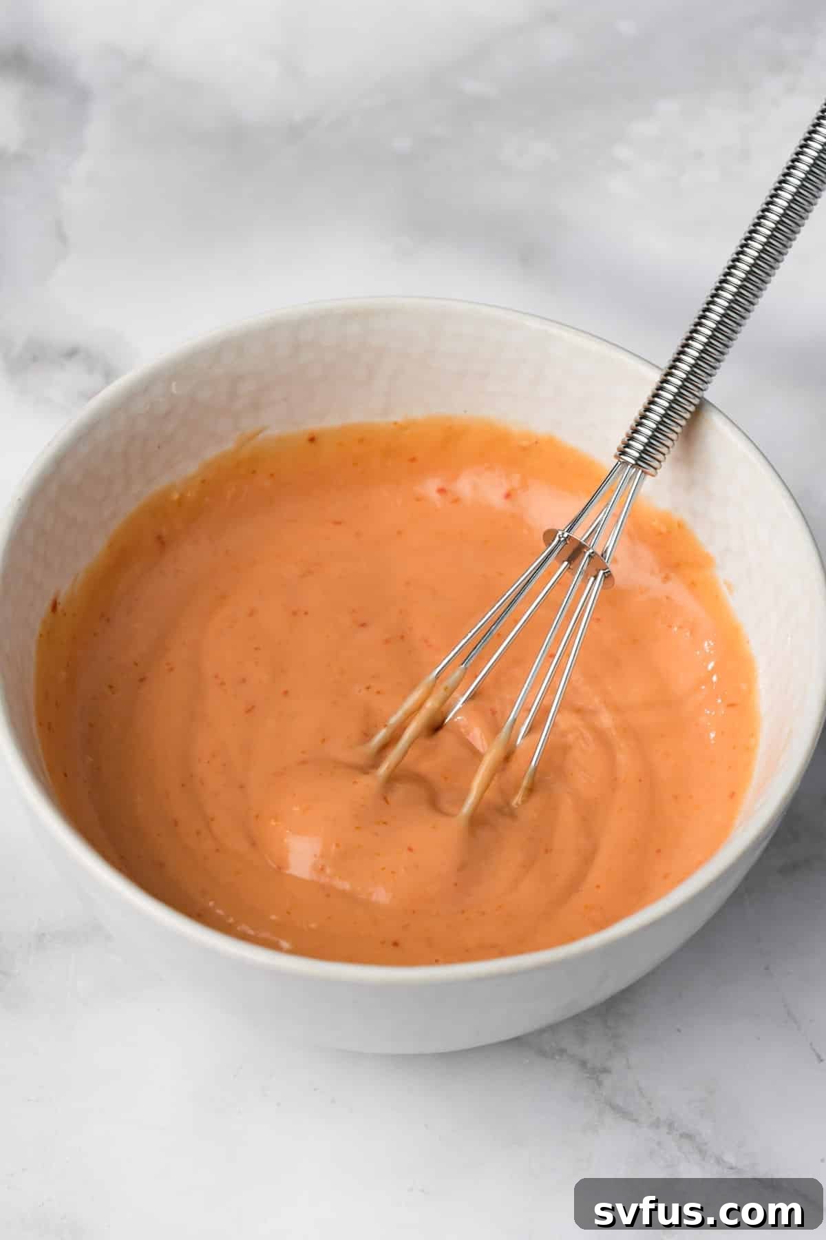 A whisk rotating in a bowl of creamy, pink-hued homemade bang bang sauce, illustrating its preparation.
