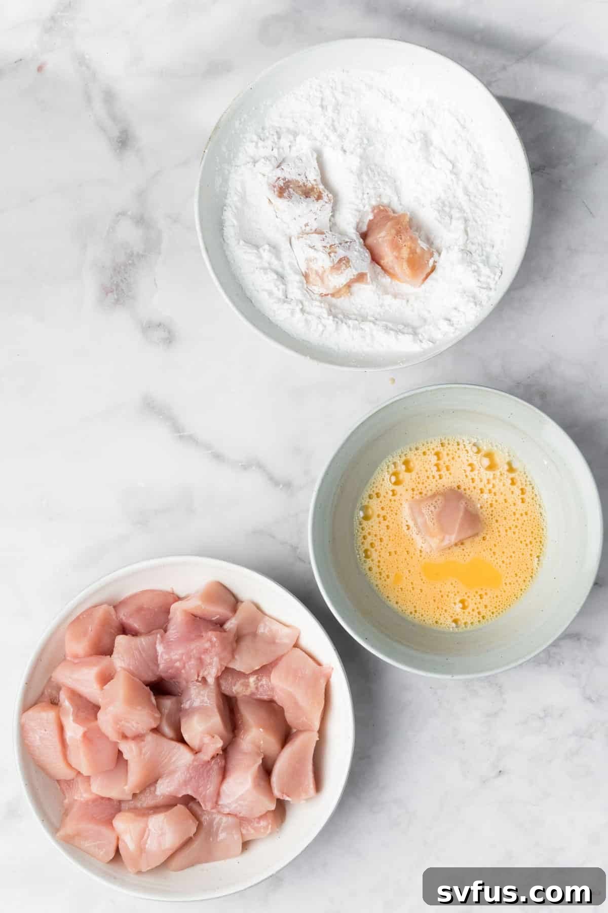 An overhead shot of raw chicken pieces, a bowl of whisked eggs, and a bowl of tapioca flour, showing the dredging process.