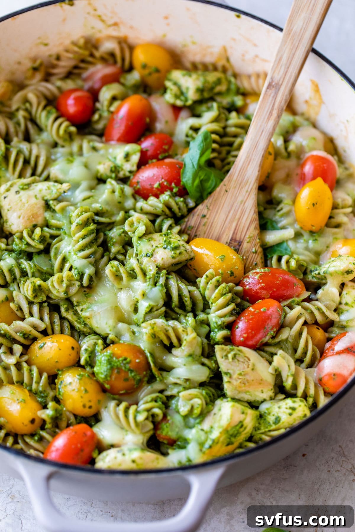A rustic skillet filled with vibrant green pesto pasta, mixed with tender chicken pieces and bright red grape tomatoes.