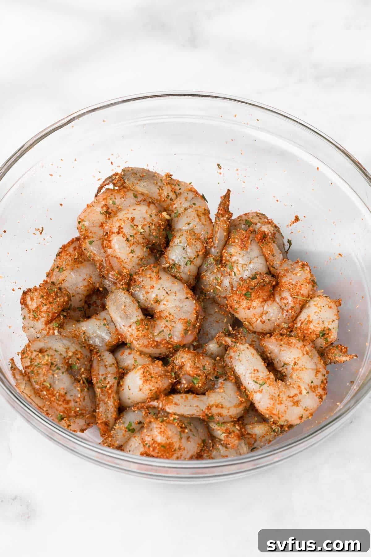 Overhead view of raw, large shrimp coated in a vibrant spice blend, resting in a clear glass bowl, ready for cooking.