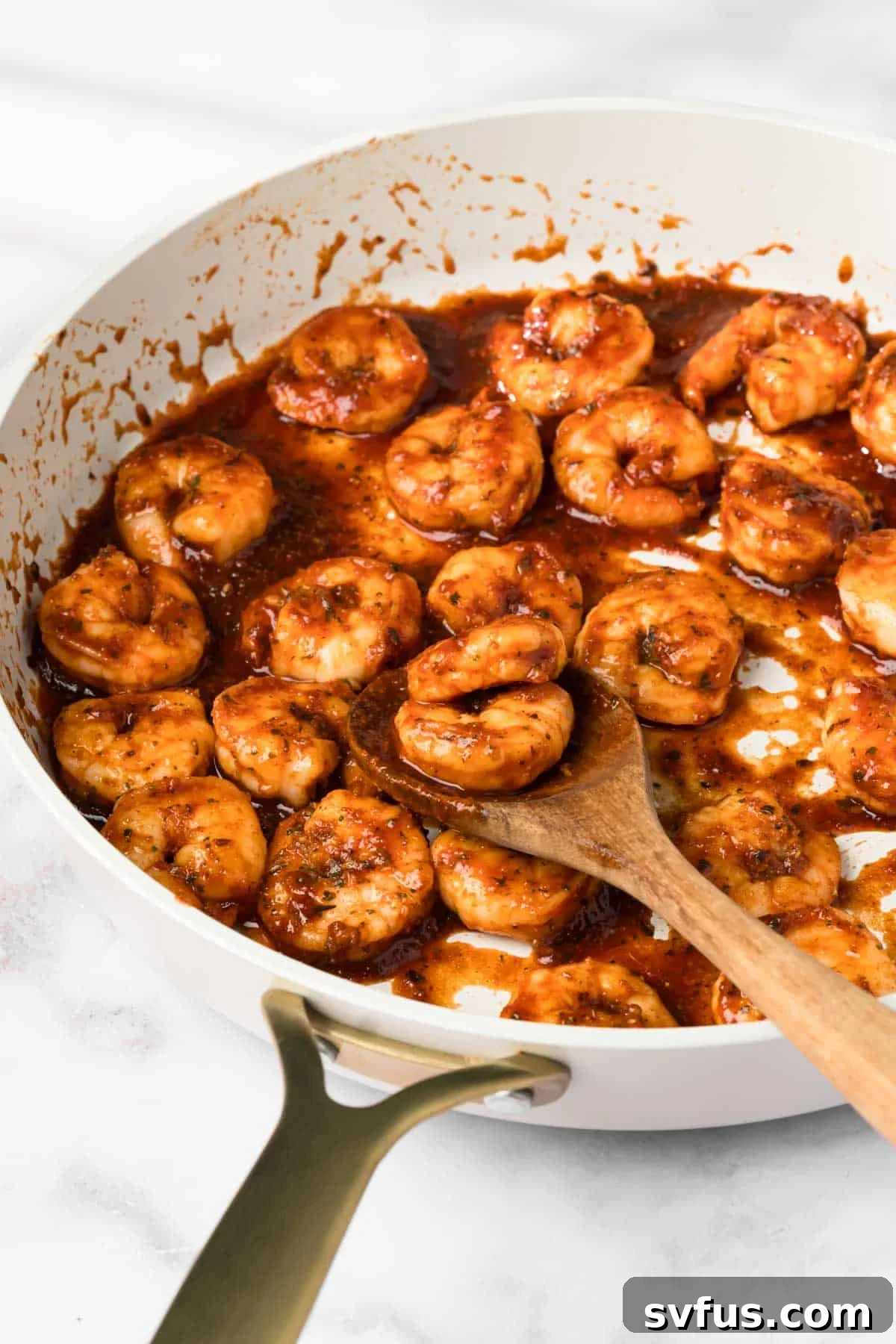 Side view of perfectly cooked BBQ shrimp glistening with sauce in a skillet, with a wooden spoon mixing them.
