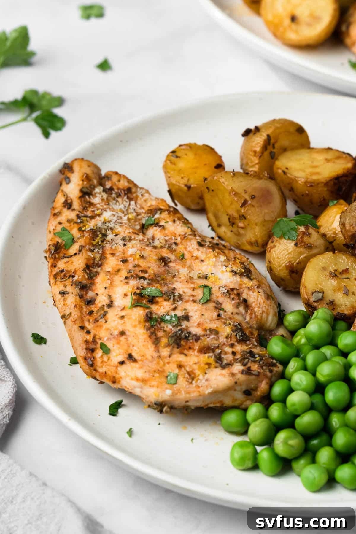 Side view of a perfectly roasted chicken breast artfully presented on a white plate, accompanied by tender roasted potatoes and bright green peas, garnished with a fresh lemon wedge.