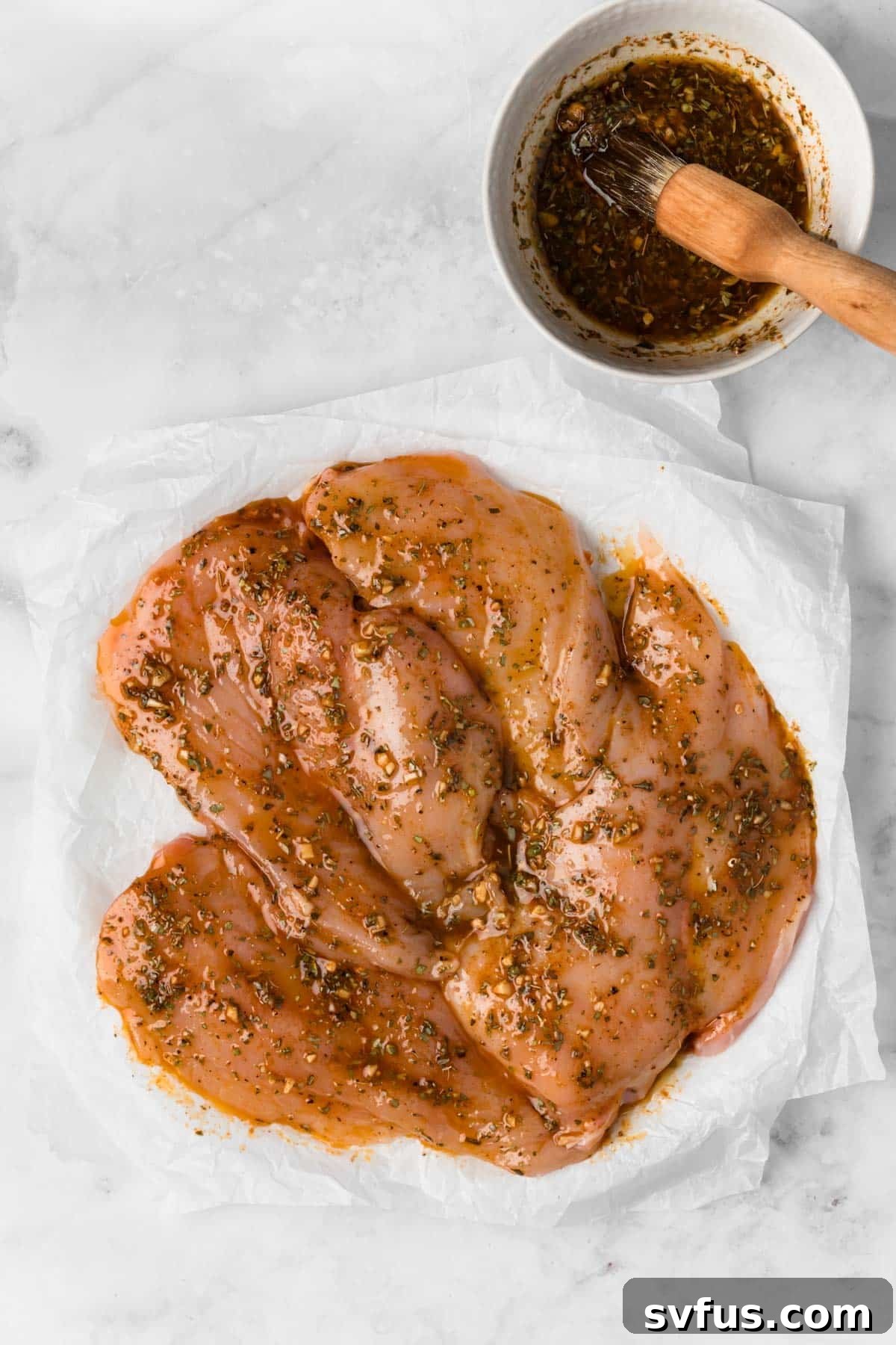 Overhead view of raw chicken breasts laid out on parchment paper, positioned next to a small bowl of the bright lemon-garlic marinade, ready for coating.