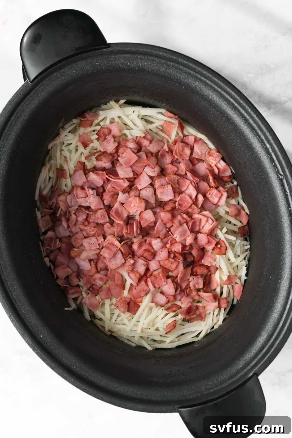 Overhead view of crispy cooked bacon layered on top of frozen hashbrowns in a slow cooker, ready for the next ingredient.