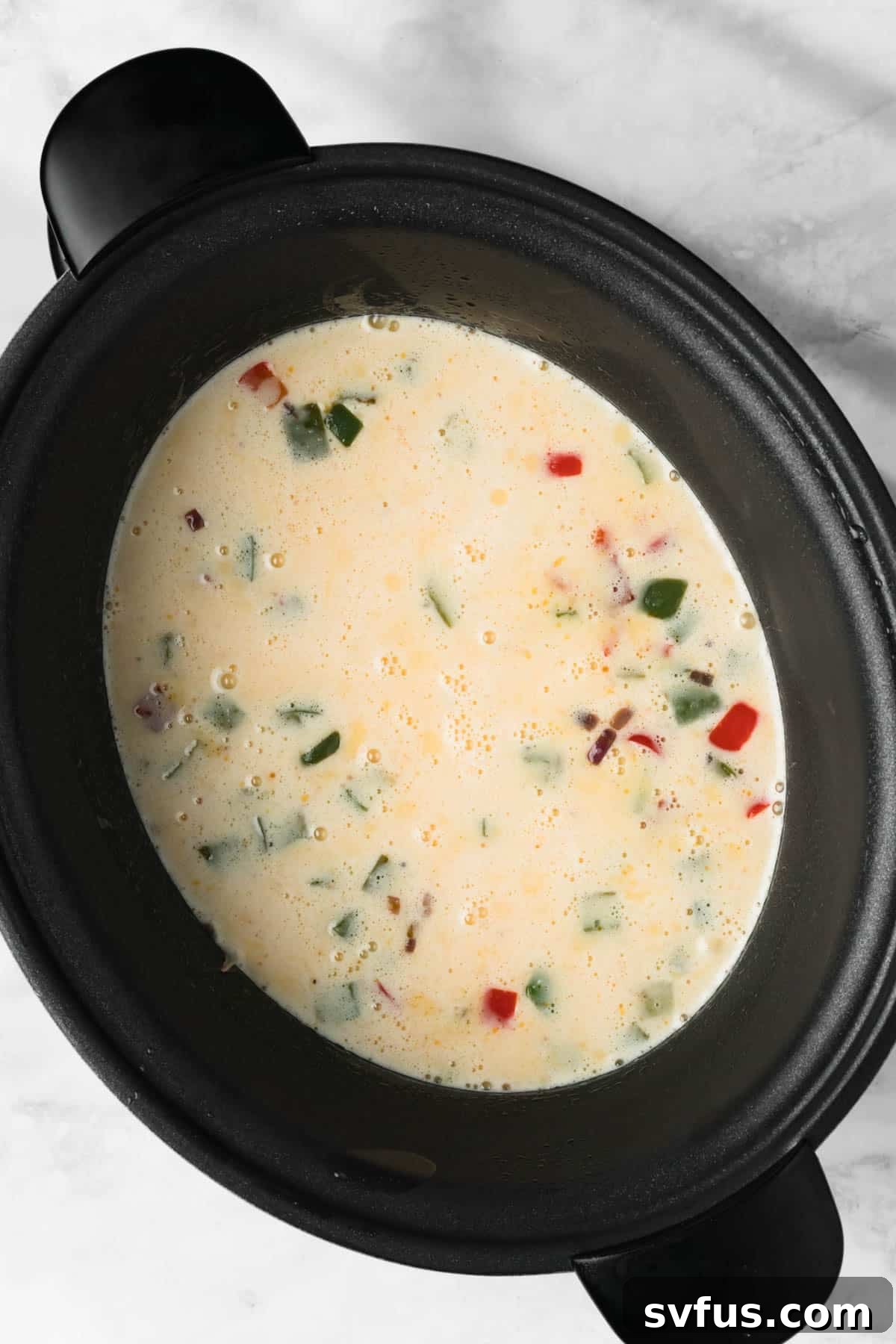 Overhead view of the liquid egg mixture poured into the crockpot, covering the hashbrowns, bacon, and vegetables, before the cheese is added.