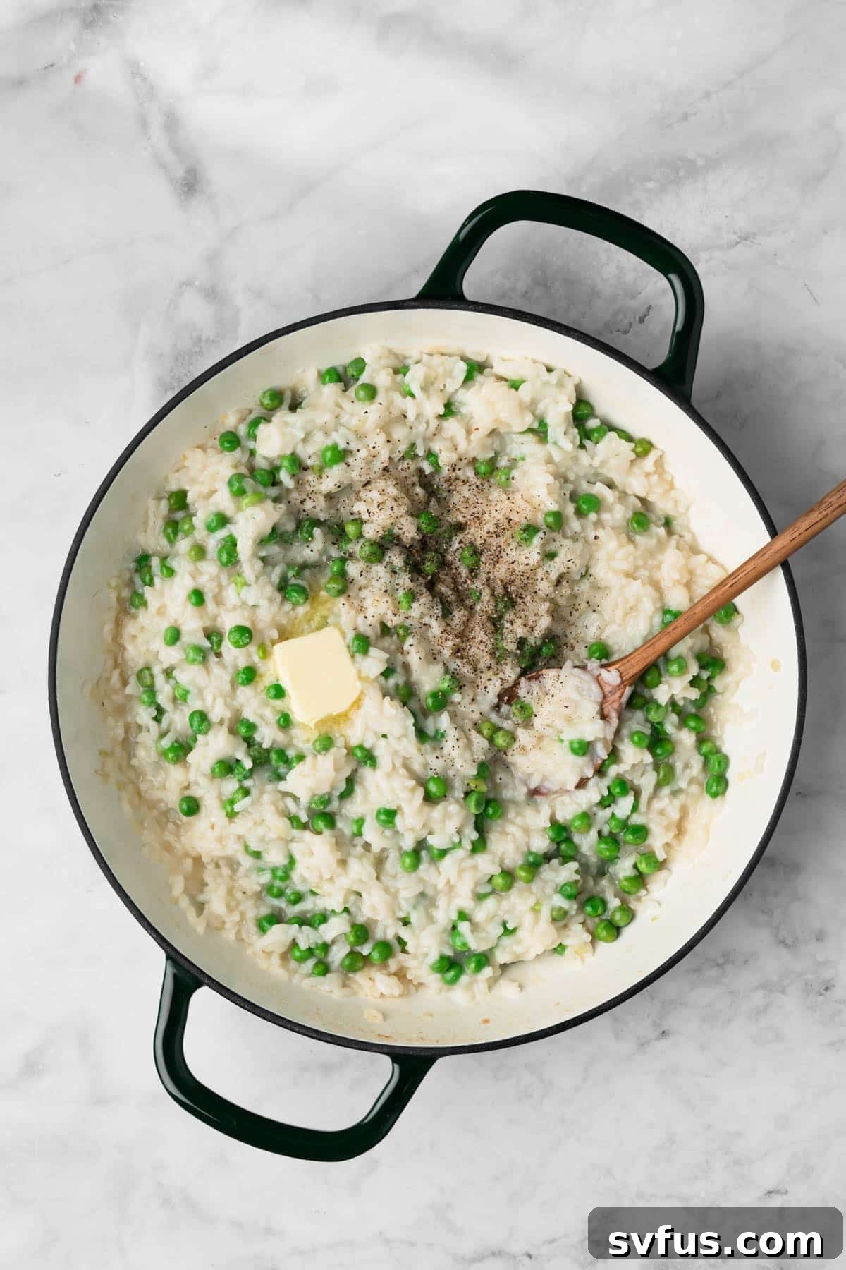 Overhead view of a wooden spoon gently stirring fresh butter, lemon juice, peas, and seasonings into a pot of finished baked risotto.