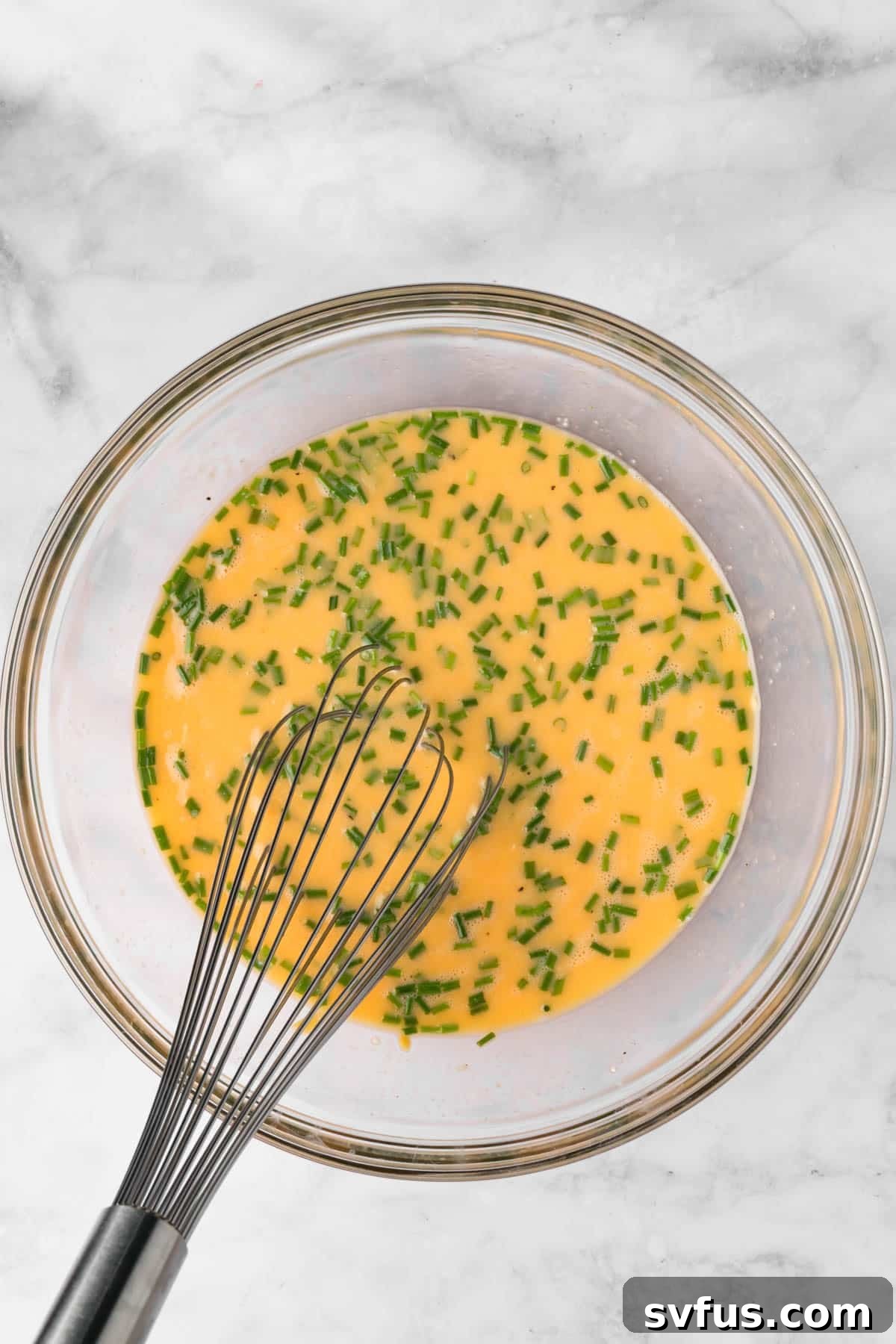 Overhead view of a whisk in a bowl with liquid eggs with chives.