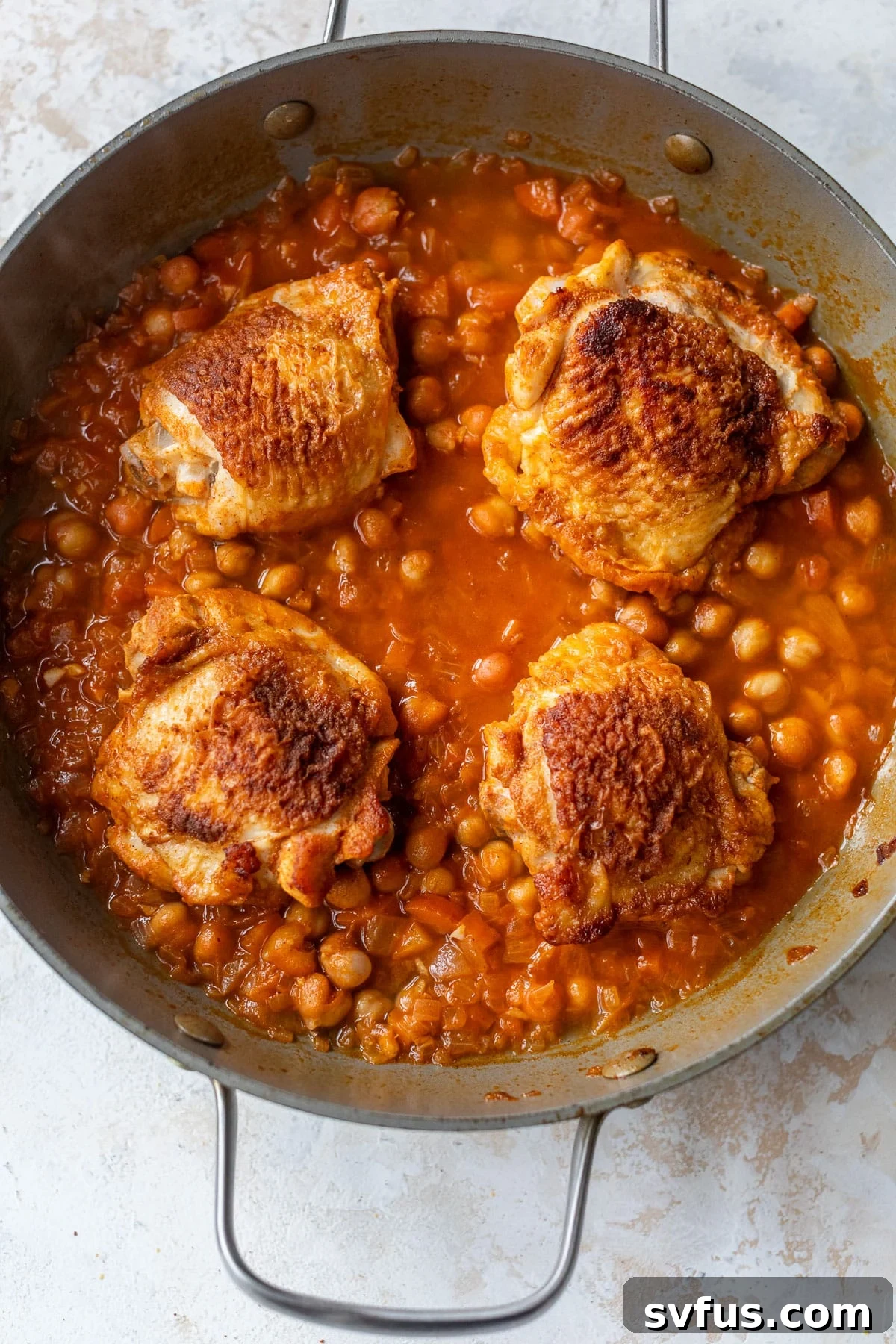 overhead view of seared chicken thighs braising in a red chickpea sauce in a skillet.